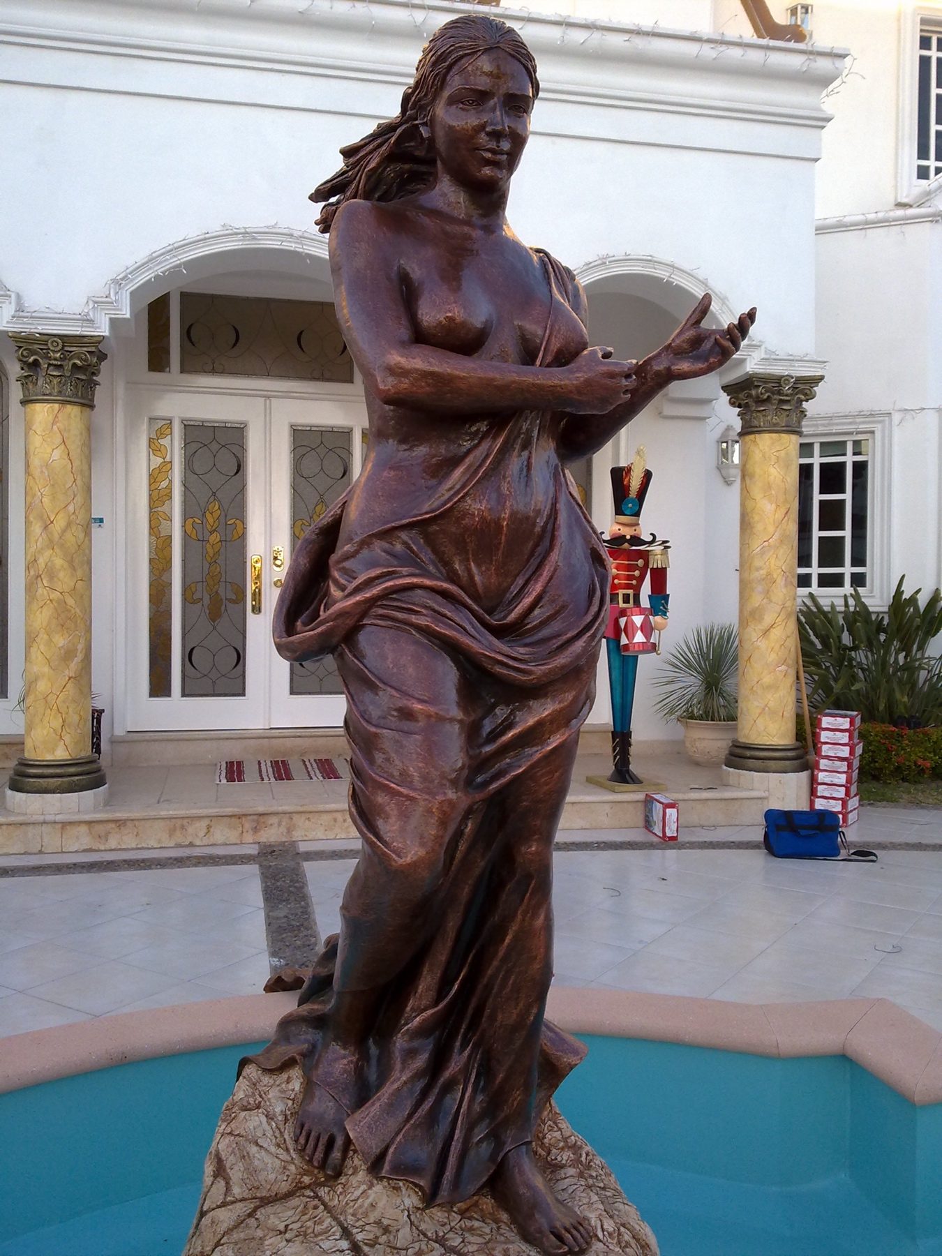 Hand-sculpted figure holding a vessel, finished in a warm bronze tone, beside a residential pool